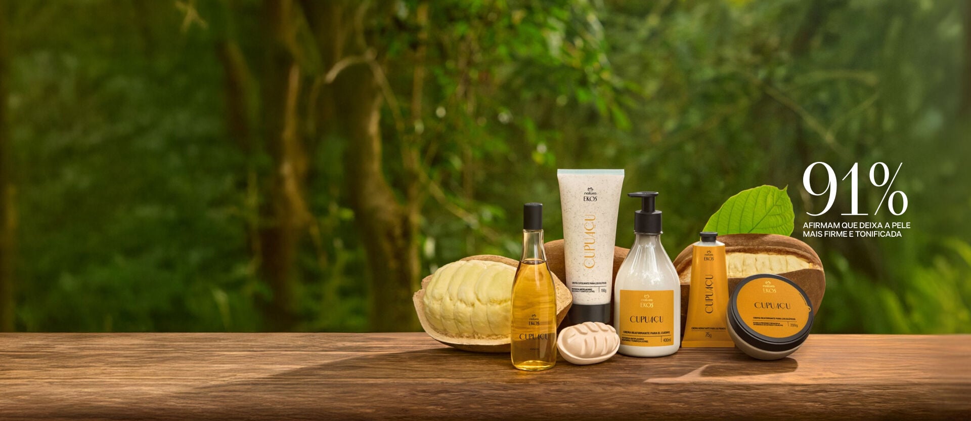 Itens Ekos Cupuaçu: bisnaga de Esfoliante, pote de Creme, frasco de Óleo Corporal e de Loção Corporal. Fundo com fruta de cupuaçu cortadas, com flores vermelhas e folhas verdes desfocadas.