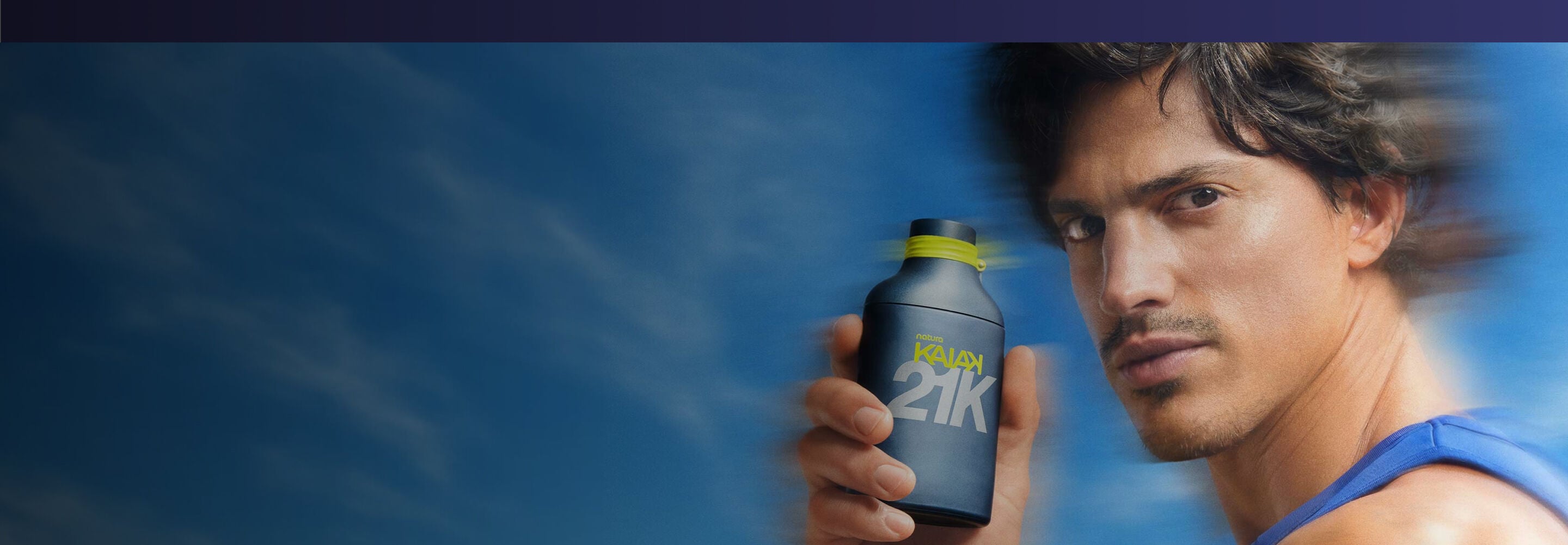 Imagem com fundo em movimento borrado de árvores verdes sob um céu azul limpo. No canto direito, há dois frascos de perfume da marca Natura Kaiak 21K posicionados lado a lado. O frasco da esquerda é mais baixo, largo e possui a cor azul-escuro fosco. O frasco da direita é mais alto, fino e de cor roxa fosca. Ambos têm detalhes em amarelo-limão na tampa e trazem o logotipo "natura KAIAK 21K" .