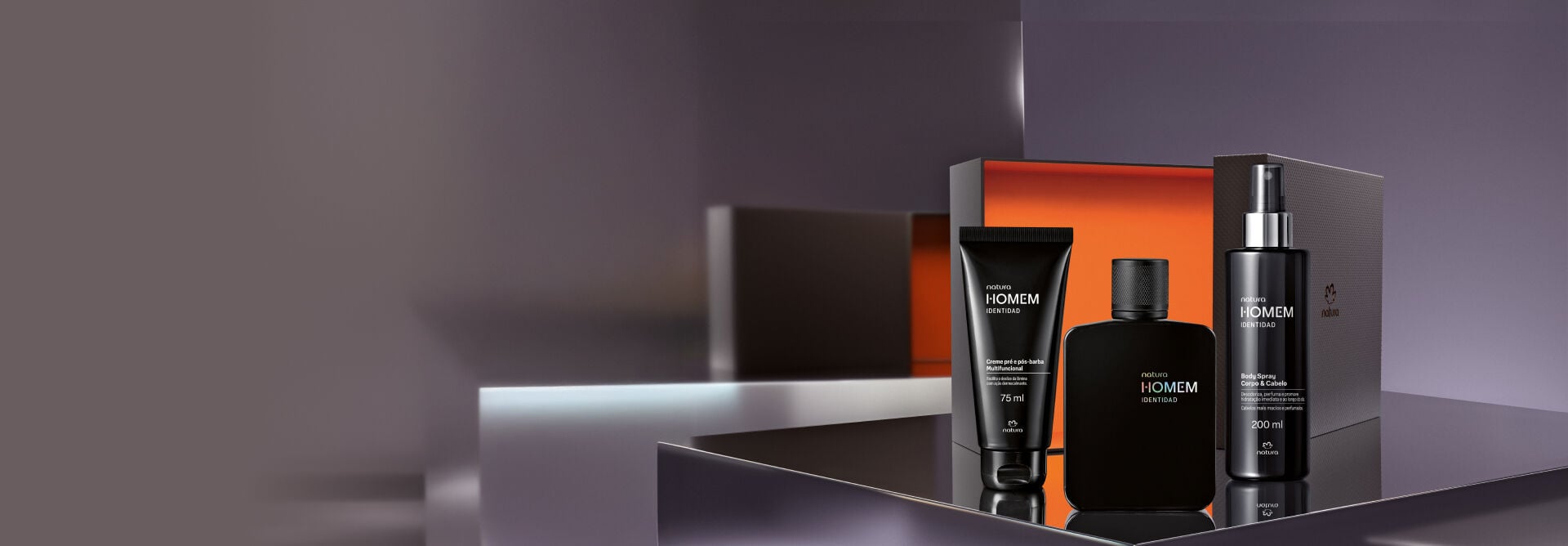 Kit Natura Homem Identidad composto por creme pré e pós-barba, colônia em frasco preto fosco e body spray para corpo e cabelo, posicionados à frente de uma embalagem de presente aberta com fundo laranja.