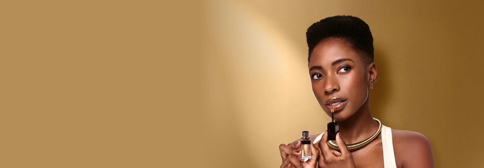 Mulher negra de cabelos curtos usa regata clara e colar dourado, segura Batom Una e aplica produto nos lábios. Fundo iluminado dourado.