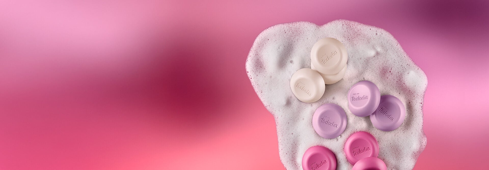 Diversos sabonetes nas cores branco, lilás e rosa sobre espuma cremosa branca. Fundo em tons de rosa.