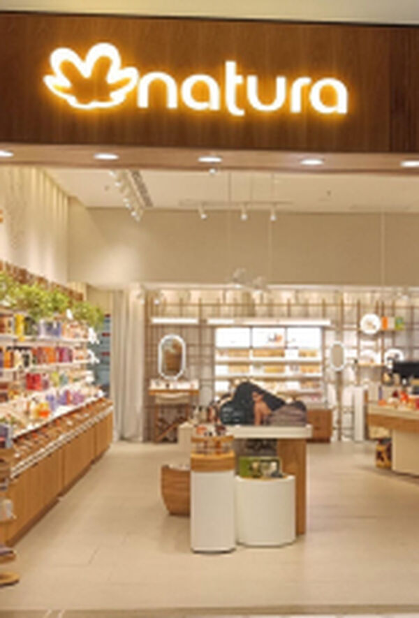 Imagem mostra uma loja física da Natura. Na entrada, logo da Natura iluminado sobre fachada de madeira e vitrine decorada. Dentro da loja, produtos organizados por categorias.