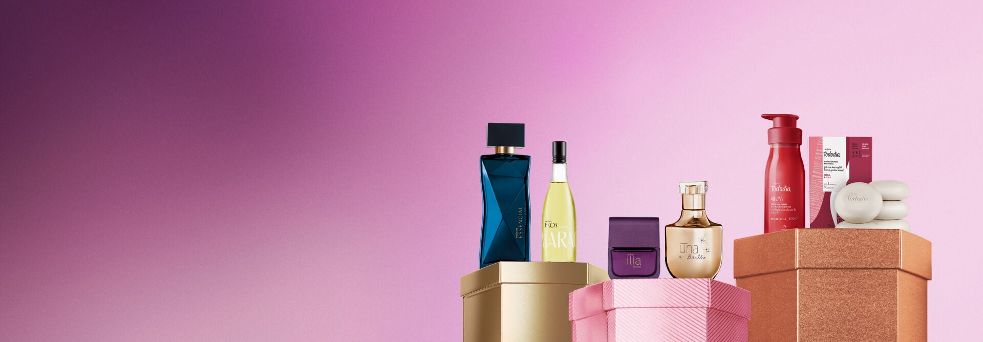 Embalagens sobre caixas brilhantes dourada, rosa e marrom, em fundo rosa: frasco de frescor Ekos Maracujá, fragrâncias Essencial Oud, Ilía Secreto e Una Brilho, e loção e sabonetes com caixa Cereja e Avelã.