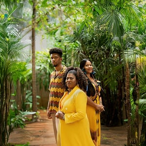 Três pessoas: uma mulher negra à frente, uma mulher indígena à direita e homem negro à esquerda, em ambiente com plantas amazônicas diversas.