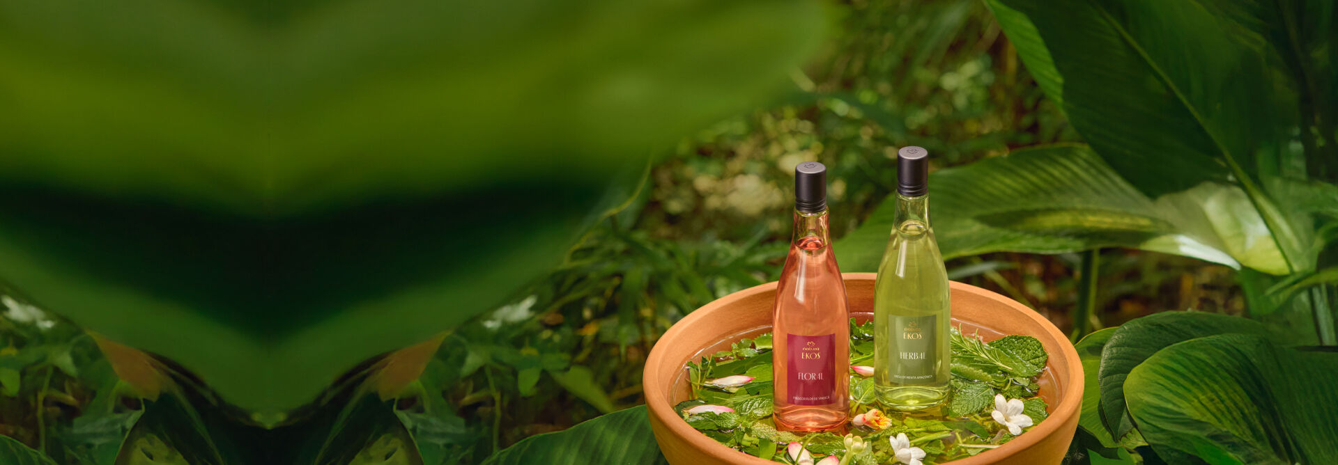 Ekos Frescores Flor de Vindicá e Hortelã Pimenta, com frascos de vidro, tampas metálicas e conteúdos rosados e verde-claros, sobre uma bacia de cerâmica com um banho de cheiro em meio à floresta. 