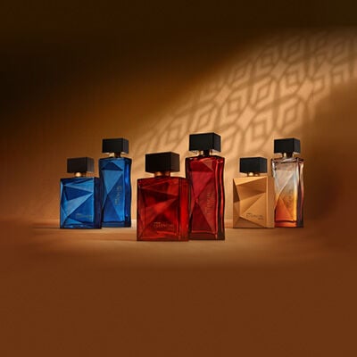 Frascos de fragrâncias Essencial masculinas e femininas: Oud em azul, Supreme em vermelho, Essencial em âmbar, sobre superfície marrom, com sombra geométrica na parede.