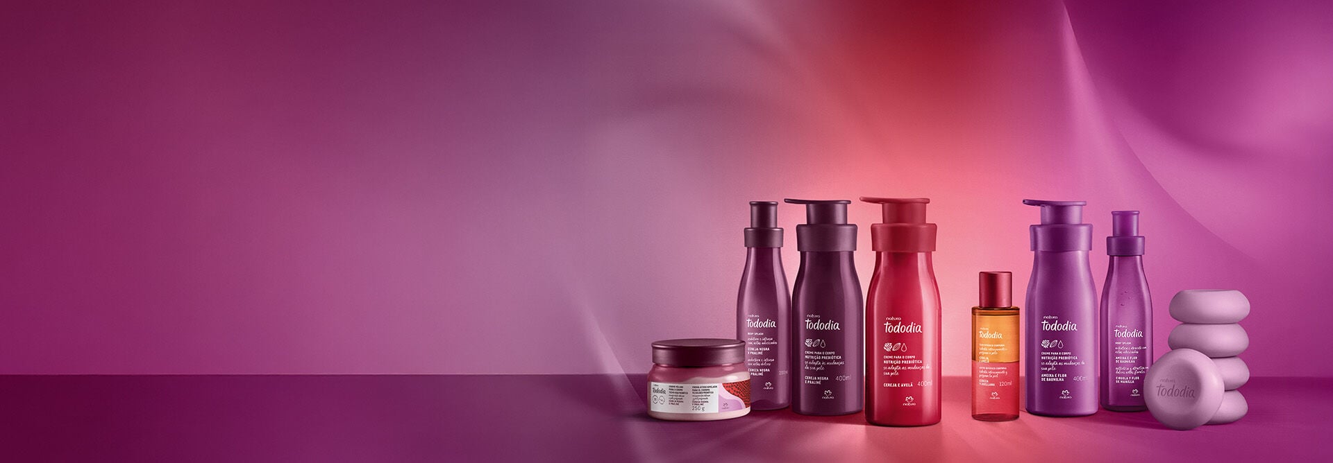 8 produtos variados de Natura Tododia nas fragrâncias Cereja Negra e Praliné, Cereja e Avelã e Ameixa e Flor de Baunilha. Embalagens roxas e vermelhas em fundo gradiente roxo, vermelho e rosa.
