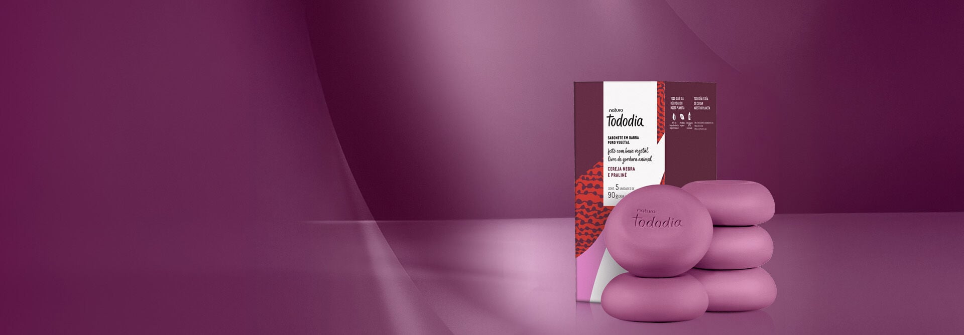 Sabonete em Barra Puro Vegetal Tododia Cereja Negra e Praliné contendo 5 barras redondas rosa empilhadas ao lado de uma caixa roxa com detalhes em vermelho, rosa e branco. Fundo roxo iluminado.