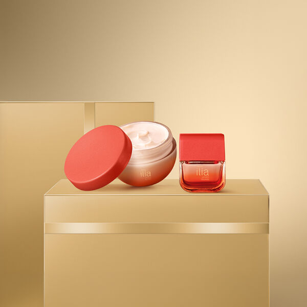 Pote de creme corporal Natura Ilía Jardim Secreto aberto mostrando textura cremosa com tampa vermelha apoiada. Ao lado, o embalagem da fragrância em tom degradê avermelhado sobre base dourada.