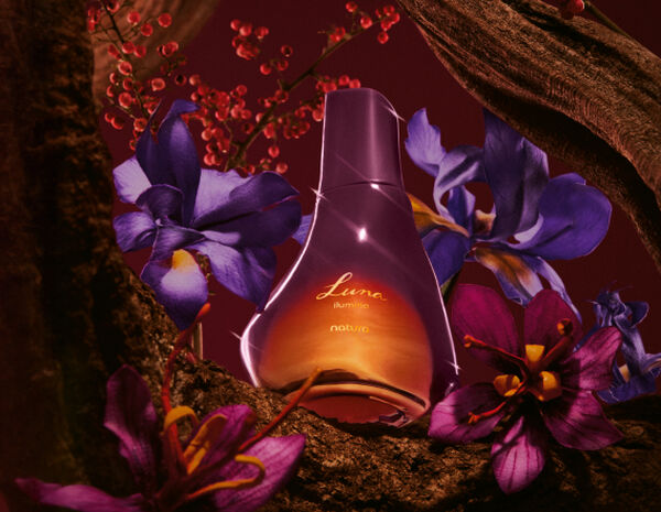 Frasco de Deo Parfum Luna Ilumina Feminino em tons de rosa e laranja sobre fundo vinho, flores violetas, troncos e texturas de madeira.