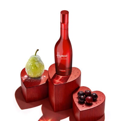 Desodorante Colônia Humor Primeiro em frasco vermelho. Ele está sobre um palco em formato de corações ao lado de frutas vermelhas redondas e uma pera verde com cristais de gelo, em fundo branco.
