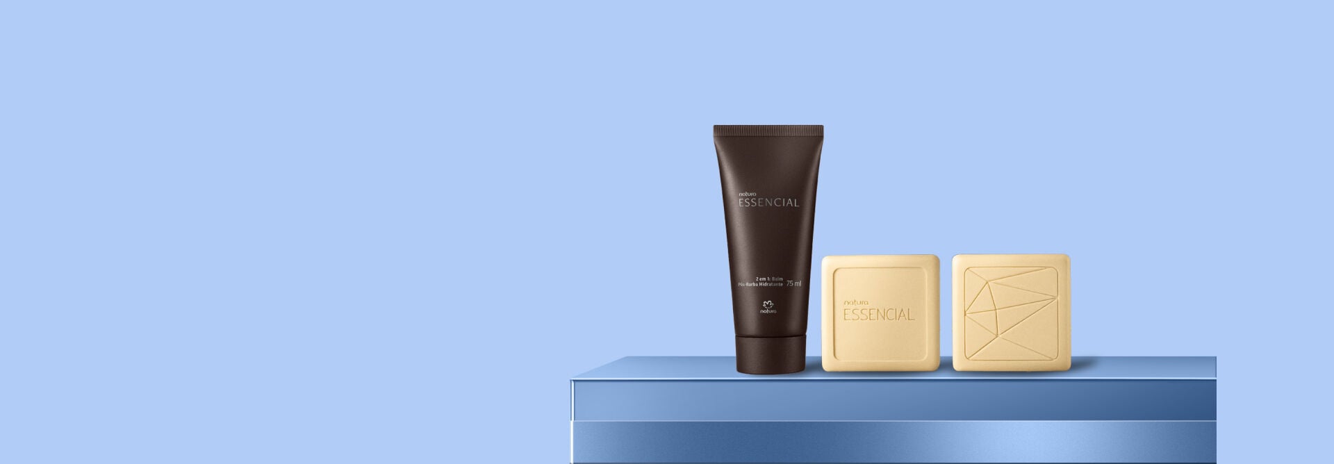 Três produtos Natura Essencial organizados sobre base azul. Uma bisnaga marrom de balm pós-barba se posiciona ao lado de dois sabonetes em barra beges, sendo um com desenho geométrica.