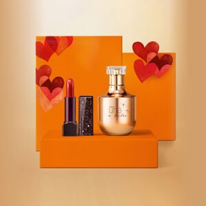 Batom Natura Una e frasco dourado da fragrância Natura Una Brilho sobre caixas de presente laranja com corações vermelhos. Fundo amarelo iluminado.