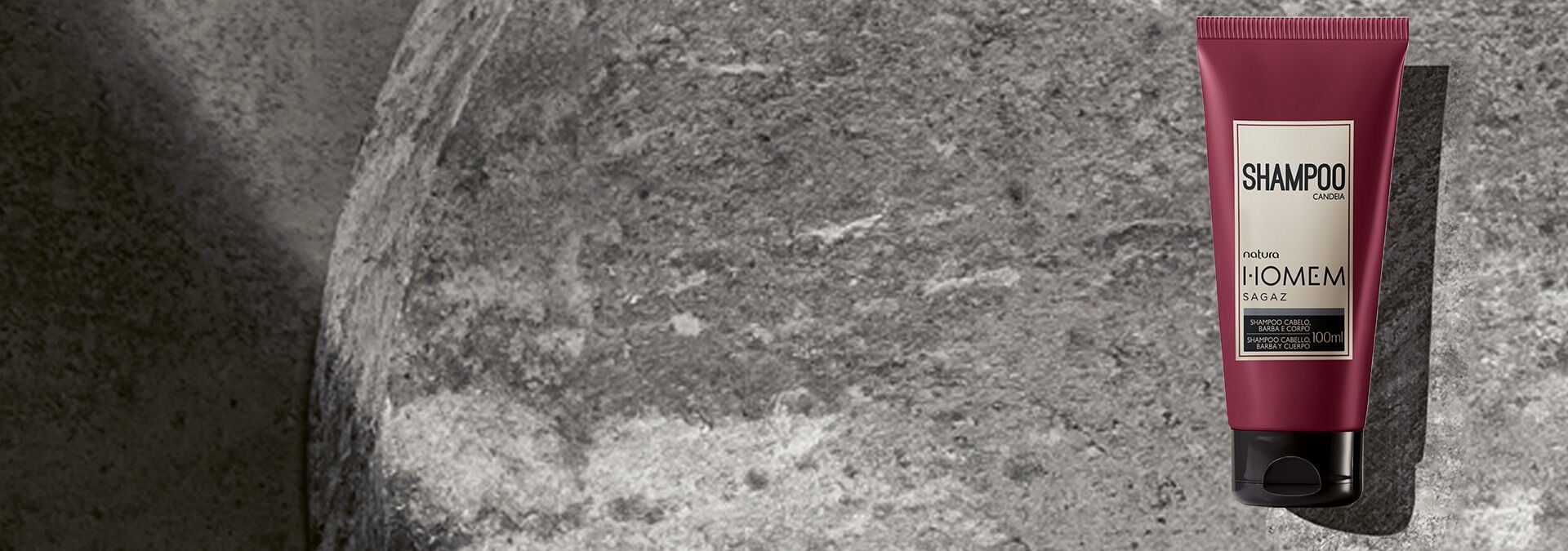 Fondo gris oscuro con una rosácea en el lado derecho, y frente a la rosácea, dos champús de la línea Homem de Natura.