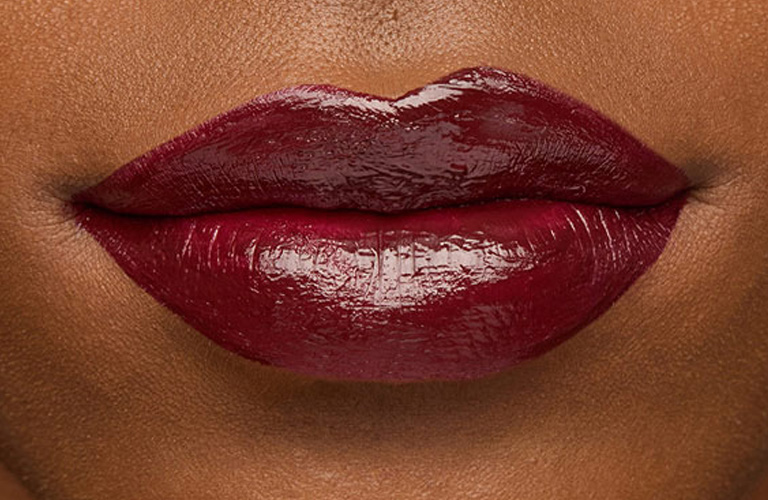 Close-up nos lábios de uma pessoa usando batom vinho escuro com acabamento vinil. A pele é morena e os lábios estão fechados.