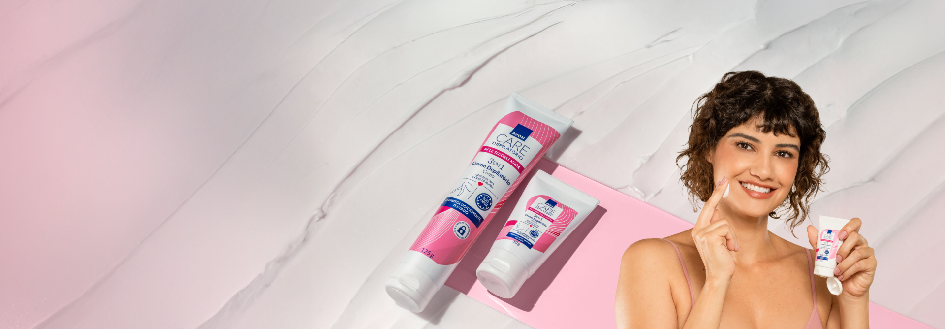 Duas bisnagas brancas com rosa, grande e pequena, de Creme Depilatório sobre textura branca e fundo rosa. À direita, mulher branca sorri aplicando creme no rosto e segura bisnaga aberta do produto.