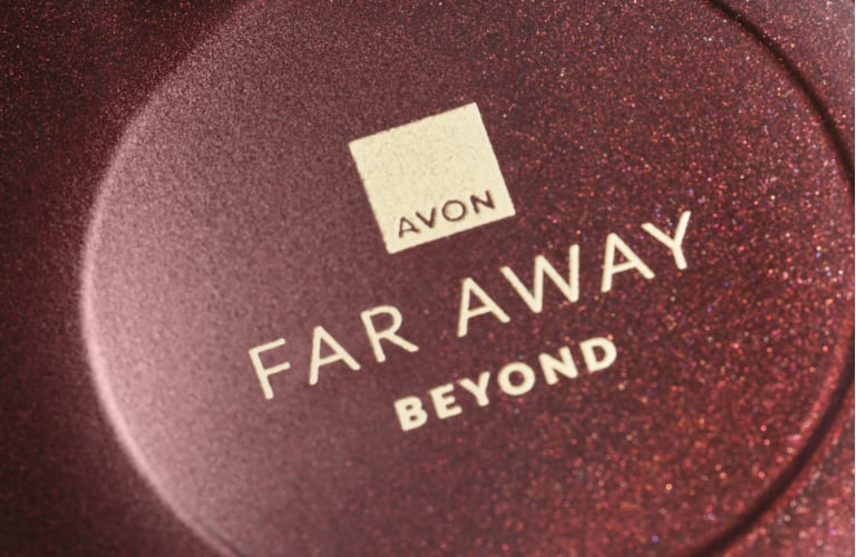 Parte superior de um frasco de perfume arredondado em tom marrom, com a válvula spray dourada voltada para o canto inferior direito. O texto no frasco está escrito "Far Away Beyond" da Avon.
