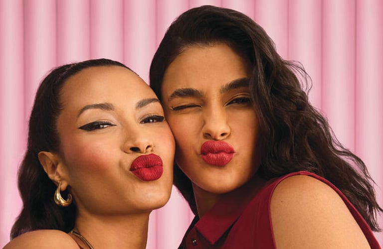Duas mulheres fazendo biquinho com boca, enquanto posam para uma foto (formato selfie). Elas vestem vestidos vermelhos, batom vermelho e tem cabelos castanhos.