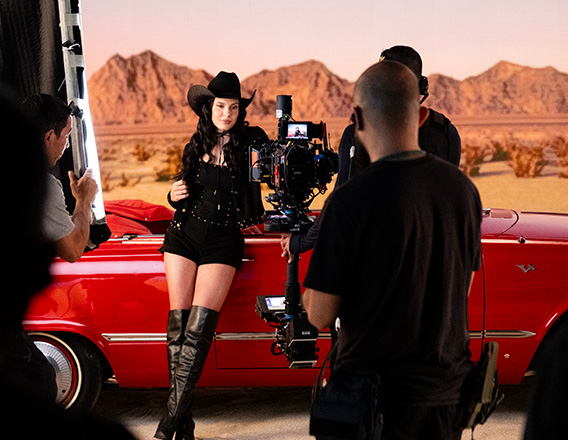 Mulher de cabelos longos castanhos, com roupa e chapéu pretos com pontos brilhantes, está à frente de carro conversível vermelho participando de gravação de vídeo. Fundo de montanhas desérticas com céu azul.