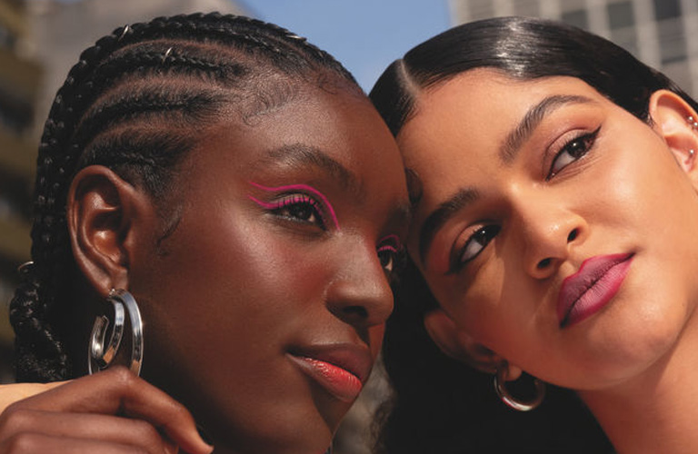 Duas mulheres posam juntas ao ar livre; à esquerda, uma mulher negra com tranças nagô usa uma jaqueta esportiva vermelha, e à direita, uma mulher de pele clara e cabelos pretos usa uma blusa verde