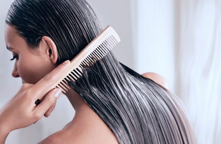 Mulher de longos cabelos pretos, lisos e sedosos. Ela está segurando uma escova de cabelo de madeira na mão direita e penteando suavemente uma mecha do cabelo para baixo.
