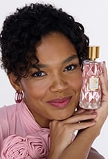 Mulher negra segura frasco da fragrância Toque de Amor Lovely com tampa dourada e líquido rosa. Ela usa roupa rosa e está em fundo claro.