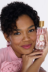 Mulher negra segura frasco da fragrância Toque de Amor Lovely com tampa dourada e líquido rosa. Ela usa roupa rosa e está em fundo claro.