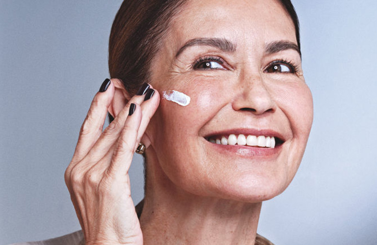 Mulher madura aplicando creme facial anti-idade Avon Renew Platinum no rosto, cuidado com a pele para firmeza e redução de rugas.