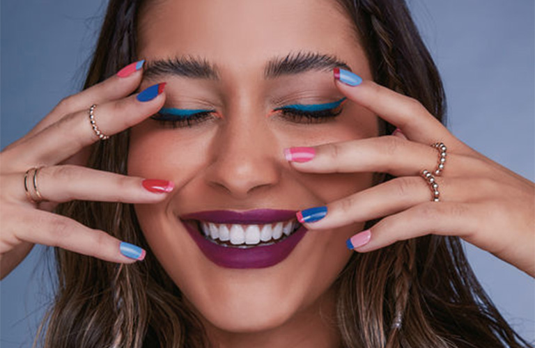 Mulher sorridente com olhos fechados, destacando delineado azul vibrante, batom roxo e unhas multicoloridas.