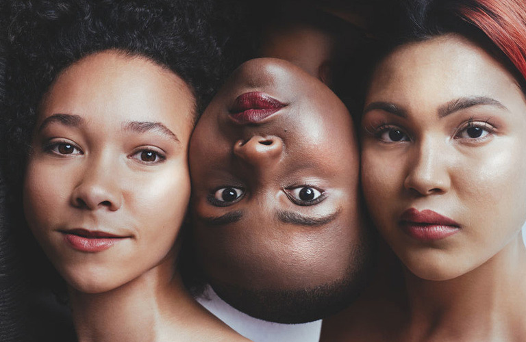 Três mulheres com diferentes tons de pele e etnias. Elas estão com maquiagem natural e a cabeça encostada uma na outra.
