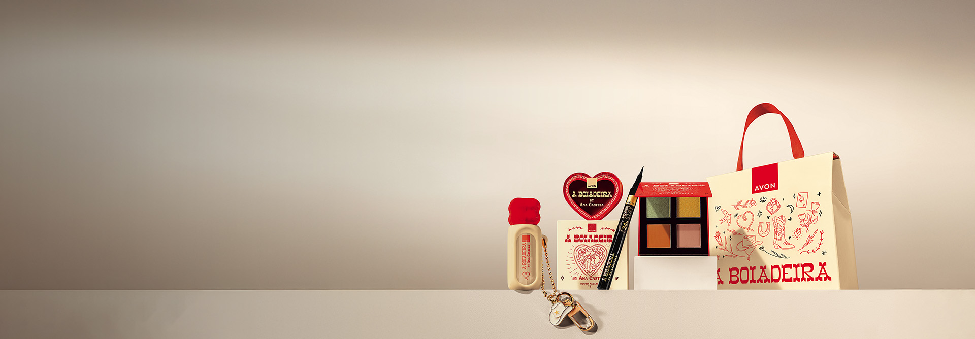 Itens da linha A Boiadeira, em bege com vermelho: Gloss Labial com pingente, Blush Facial em formato de coração com caixa, Caneta Delineadora preta, Paleta de Sombras e Sacola Especial, em fundo bege.