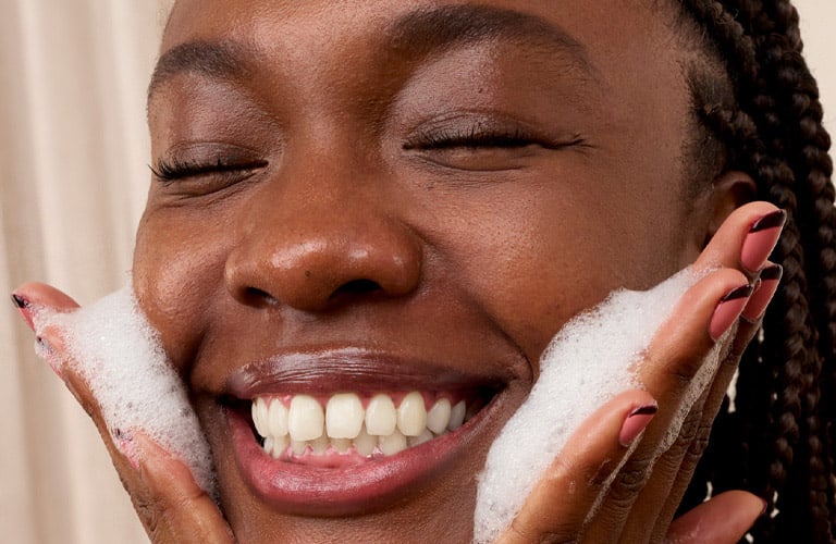 Mulher negra sorridente e com olhos fechados lava o rosto. Ela tem as mãos cobertas por uma espuma branca, encostadas nas bochechas. Seu cabelo está preso em tranças nagô e suas unhas estão pintadas em tons de rosa.
