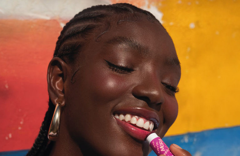 Duas mulheres com maquiagens marcantes. À esquerda, uma mulher de pele negra usa uma sombra cintilante em tons de azul e cabelo com trança nagô. À direita, uma mulher de pele parda usa sombras em tons terrosos e cabelos raspados e amarelados.
