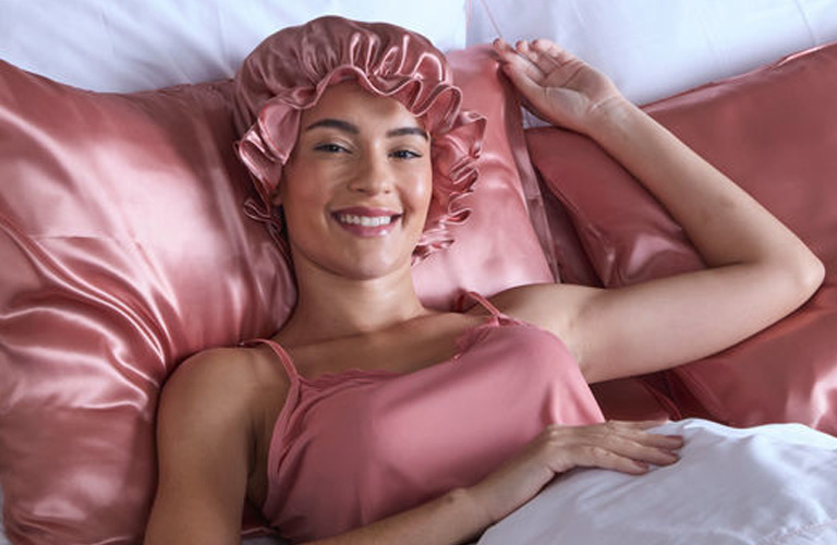 Mulher sorridente usa touca de cetim rosa para os cabelos, enquanto está deitada em uma cama com lençois brancos e fronhas de cetim na cor rosa.