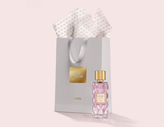 Frasco em vidro facetado e tampa dourada de Charisma Magnetic à frente de sacola de presente cinza com texto Iconic Collection Avon, e papel de seda branco. Fundo claro.