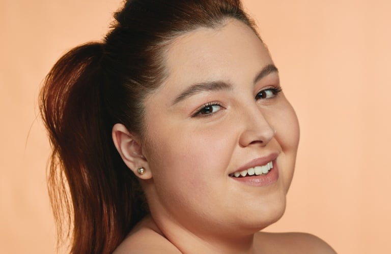 Imagem close-up em rosto de uma mulher branca sorridente, com cabelos lisos castanhos e amarrados em um rabo de cavalo frouxo. Ela está em um fundo rosa-claro de estúdio.