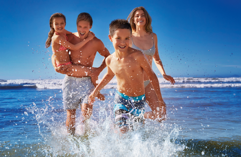 Família sorridente se divertindo à beira-mar, com um menino correndo para fora do mar e espalhando água da praia, enquanto o pai carrega a filha no colo e a mãe caminha ao fundo sob um céu azul.