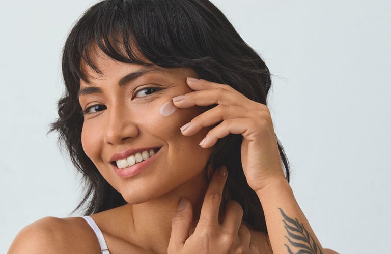 Mulher de cabelos pretos com franja, sorrindo enquanto aplica ácido glicólico na bochecha. Ela possui diversas tatuagens pretas nos braços, incluindo desenhos de flores, abelha e folhagens.