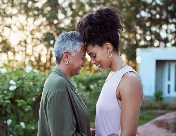 Mulheres negras de idades diferentes, frente a frente com as testas encostadas. À esquerda, mulher de cabelos curtos grisalhos de verde e, à direita, mulher de cabelos presos e crespos de rosa. Fundo externo doméstico.