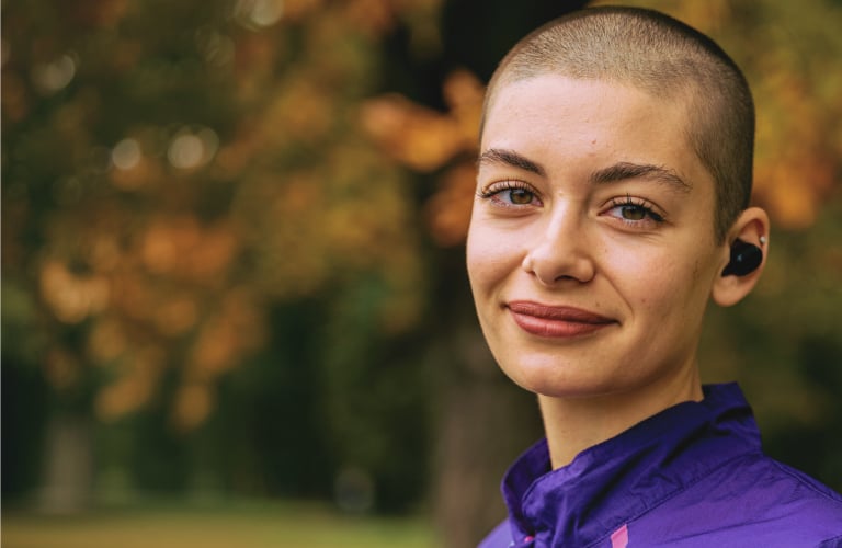 Mulher sorridente de pele clara e cabelos raspados curtos (careca) veste uma jaqueta esportiva roxa e usa fone de ouvido sem fio do tipo na cor preta. O fundo da imagem está desfocado, revelando um ambiente arborizado.