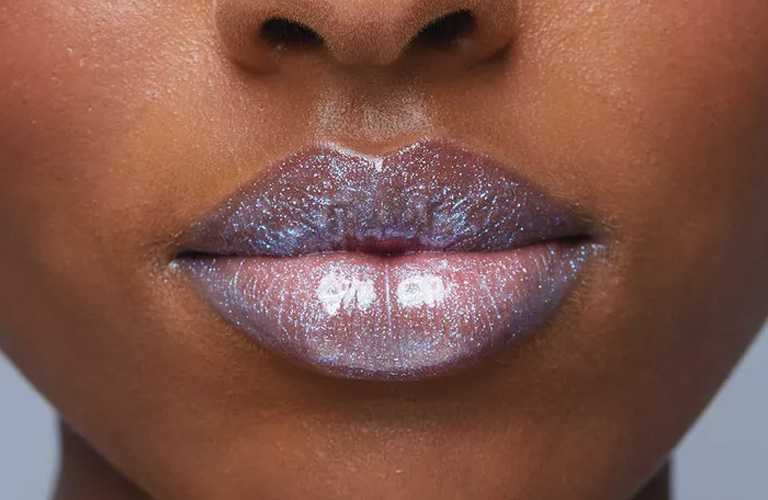 Imagem close-up nos lábios de uma mulher negra usando um gloss holográfico da marca Avon na cor azul claro para o carnaval.