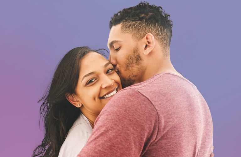 Mulher sorridente de cabelos longos castanhos está abraçando um homem de cabelos curtos que também beija o topo da cabeça desta mulher.