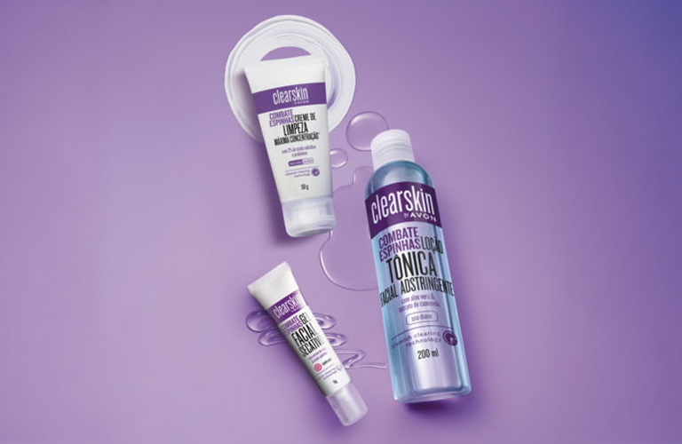 Três produtos da linha Clearskin para tipos de acne sobre um fundo roxo sólido, acompanhados por respingos transparentes e um círculo de creme branco. Os produtos incluem duas bisnagas brancas com detalhes em roxo e um frasco do tônico adstringente de acne da Avon.
