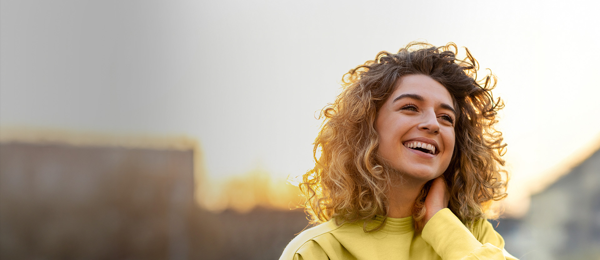 Mulher branca de cabelos castanho-claros ondulados sorridente, com blusa amarela. Fundo iluminado ensolarado.