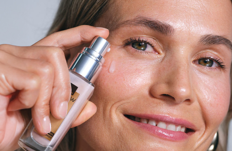 Mulher sorridente de cabelos grisalhos segura o Power Sérum Renovador da Renew próximo ao rosto, simulando o momento de aplicação.