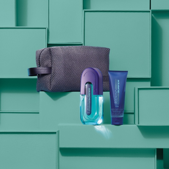 Frasco em tons de azul de Deo Colônia 300 Km/h Surfer, bisnaga azul de Shampoo para Cabelo e Corpo 300 Km/h Surfer, com nécessaire cinza com alça lateral, sobre caixas verdes, que compõem o fundo.