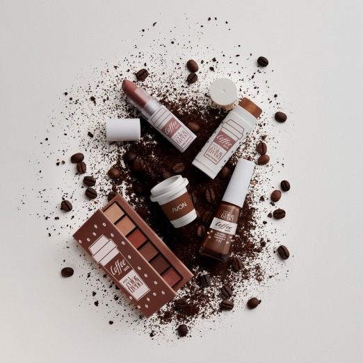 Batom e Multi Stick da linha Avon Color Trend Coffee Date mergulhando em xícara branca com conteúdo marrom leitoso. Fundo branco.