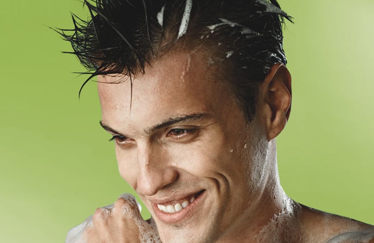 Homem sorridente de cabelo curto e espetado, toma banho com espuma de sabão, enquanto lava os cabelos.