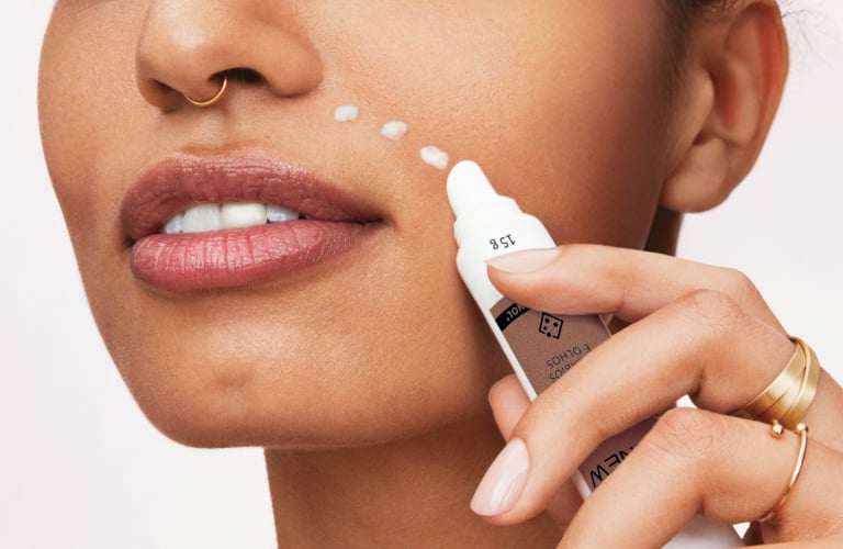 Mulher de cabelo cacheado curto aplicando um creme facial de Protinol perto dos olhos. Ela segura a ponta de uma bisnaga na região da bochecha, olhando para frente com um leve sorriso.