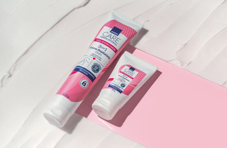Dois cremes depilatórios para o corpo em bisnagas brancas com detalhes em rosa e azul da linha Avon Care. Eles estão deitados sobre uma superfície texturizada que imita uma loção branca, com uma faixa diagonal rosa sólida servindo de base para os produtos.